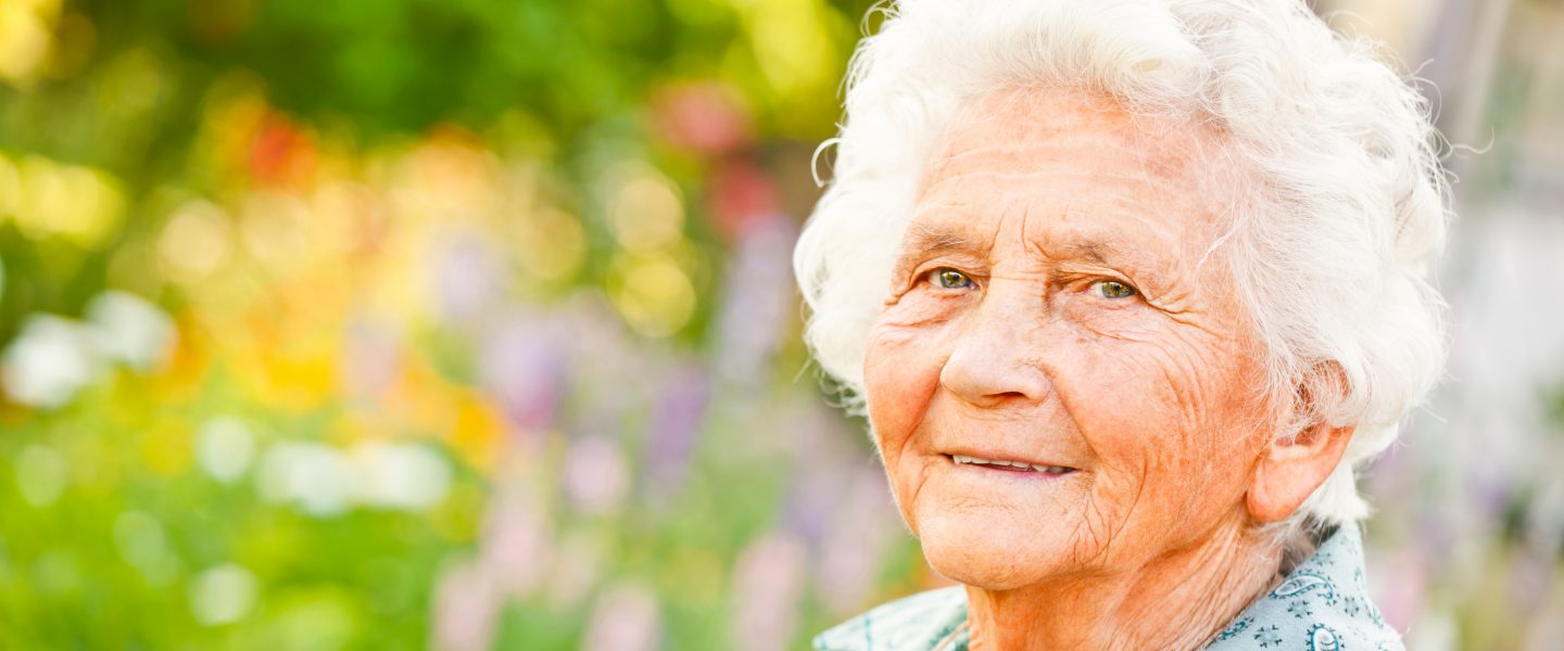 Ältere Frau mit weißem Haar lächelt vor buntem Blumenfeld im Hintergrund, strahlt Freude und Lebensweisheit aus.