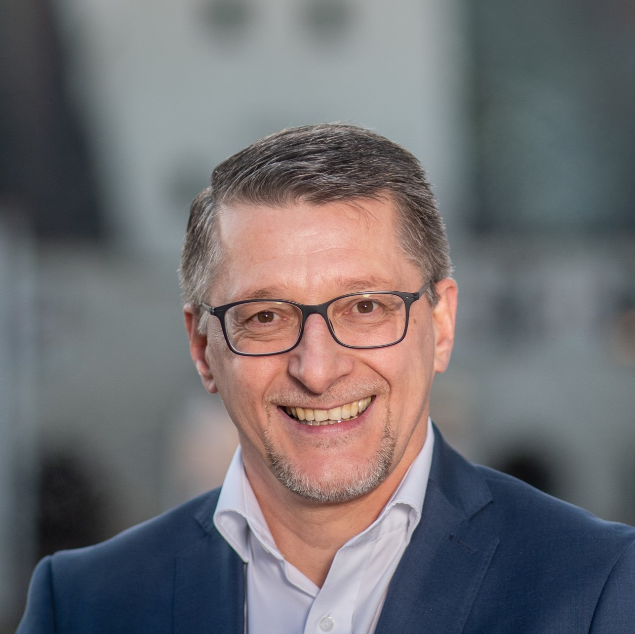 Lächelnder Mann mit Brille in Anzug, vor unscharfem Hintergrund, zeigt Selbstbewusstsein und Freundlichkeit.